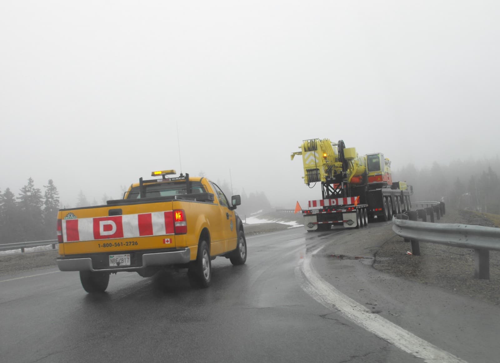 A pilot car with an oversize load sign leading a wide load along a wet highway